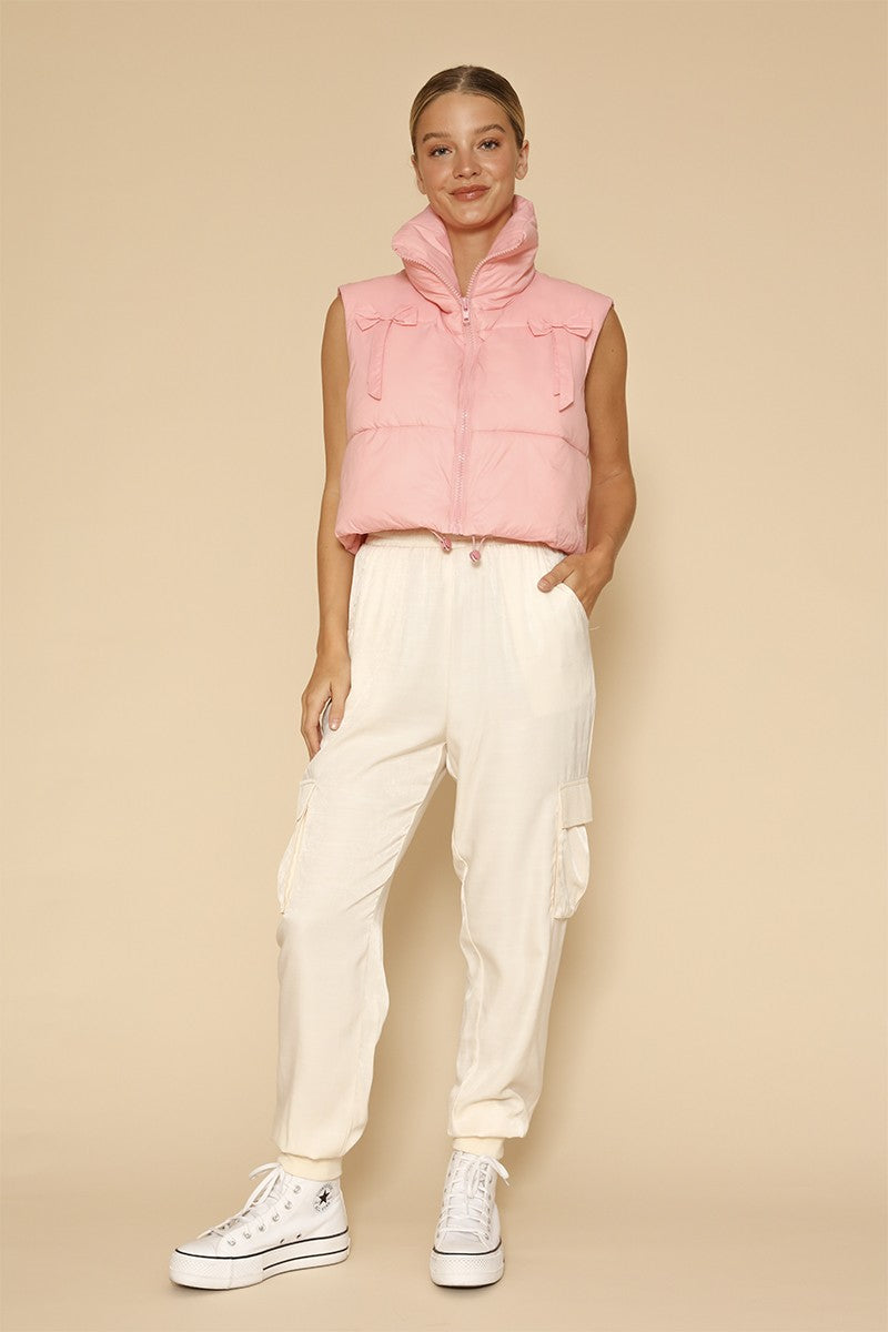 Woman wearing a pink puffer vest with bow ties and white pants on a beige background.