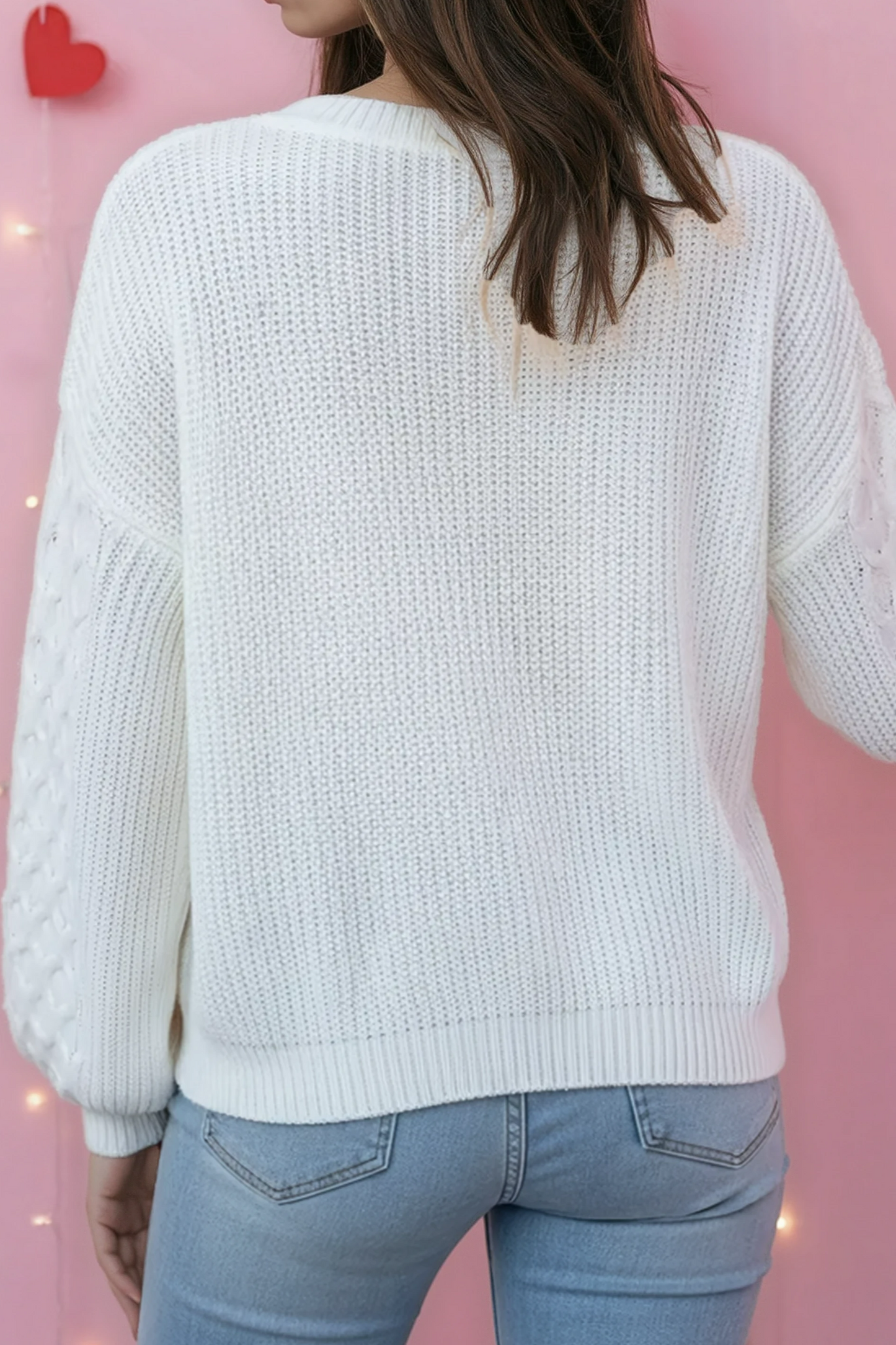 Person wearing a white sweater and light blue jeans against a pink background with heart decorations.