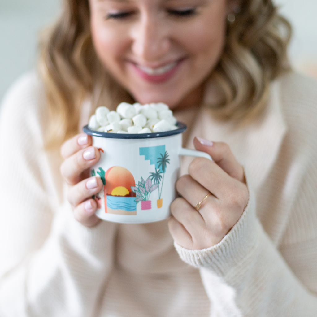 Woman holding a mug with hot chocolate and marshmallows, wearing a beige sweater.