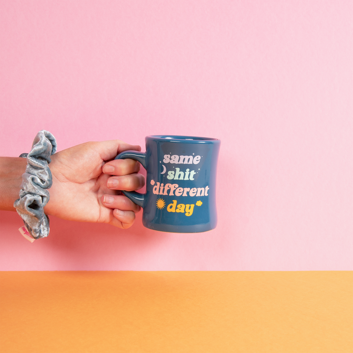 Hand holding a "same sh** different day " mug with text against a pink and orange background