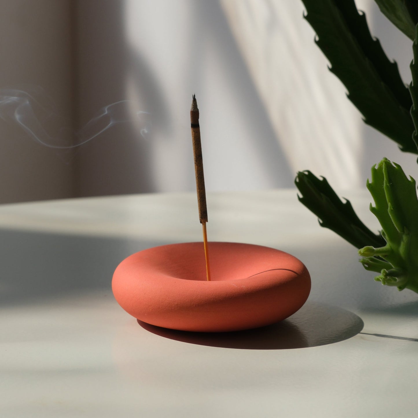 handmade Orange incense burner with a single stick of incense on a light surface, with a plant in the background.