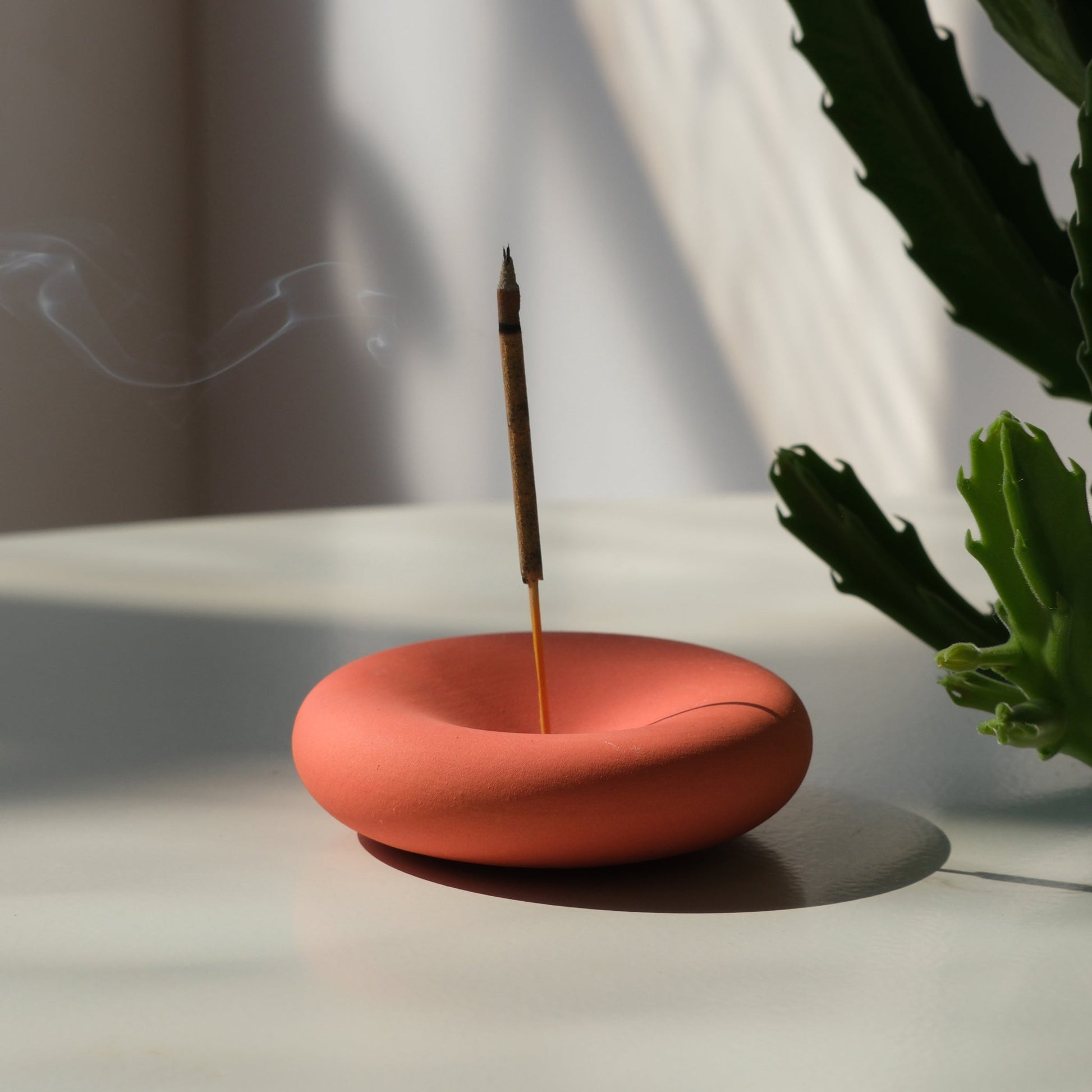 handmade Orange incense burner with a single stick of incense on a light surface, with a plant in the background.