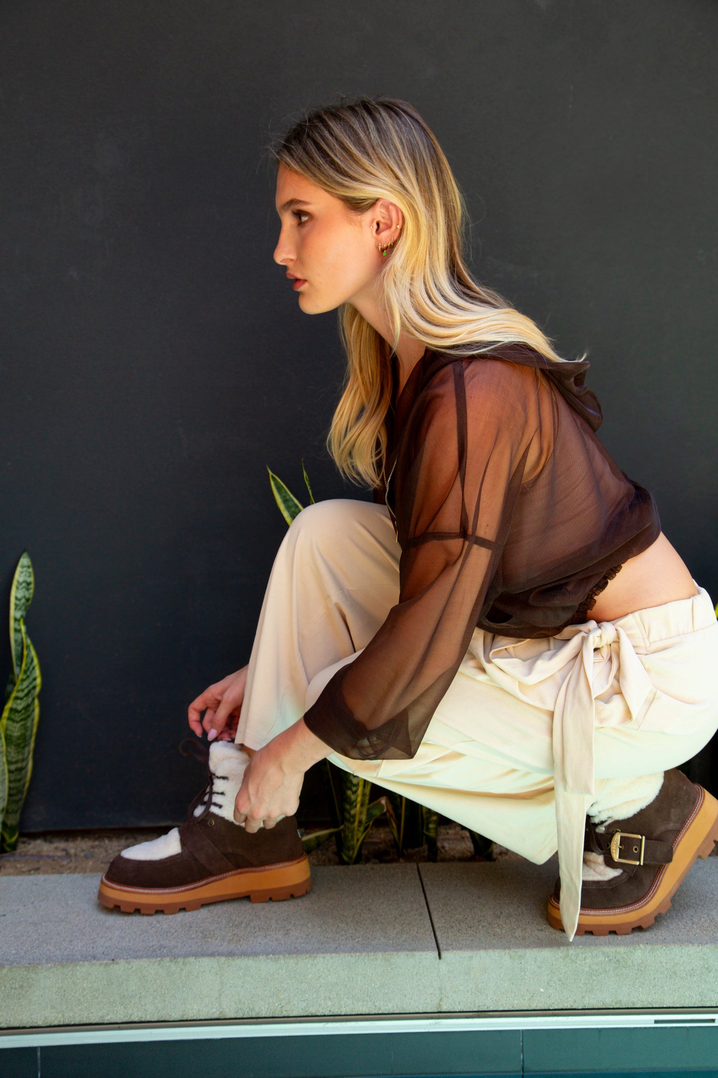 Woman sitting on a step wearing brown shoes and a sheer top with beige pants.