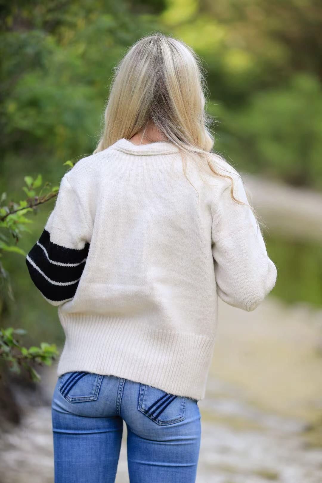 Person wearing a white sweater with black sleeves and blue jeans in a natural setting.