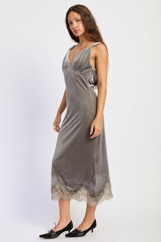 A woman modeling a sleeveless gray velvet midi slip dress with a lace-trimmed hem, paired with black pointed-toe flats, against a plain light gray studio background.