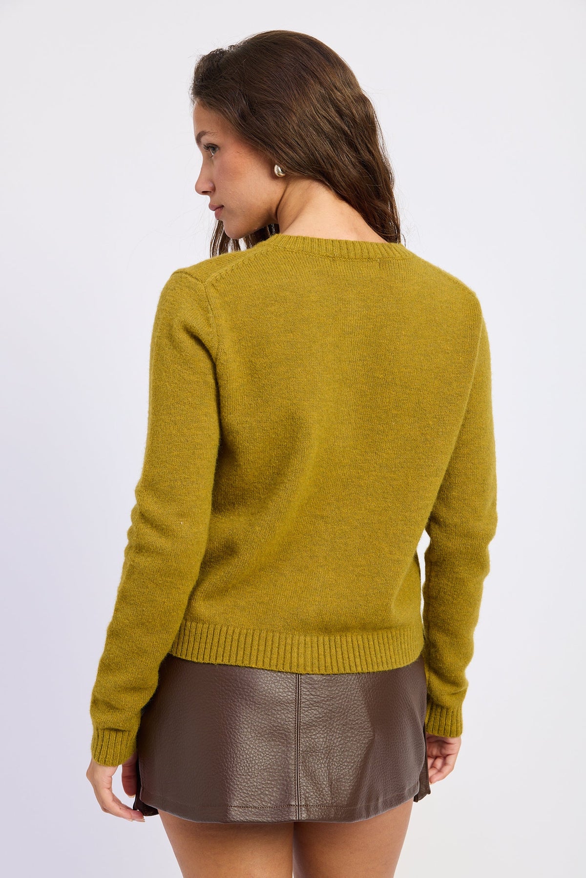 A woman shown from the back wearing a mustard yellow knit sweater and a brown faux-leather mini skirt, against a plain light gray studio background.
