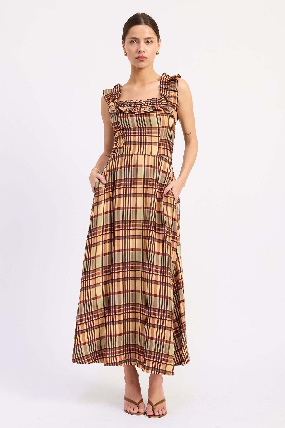 A woman modeling a sleeveless maxi dress with a ruffled neckline and a plaid pattern in beige, brown, and red, standing against a plain white studio background.