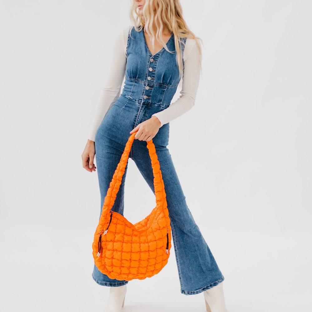 Person wearing a denim jumpsuit holding an orange quilted handbag against a white background