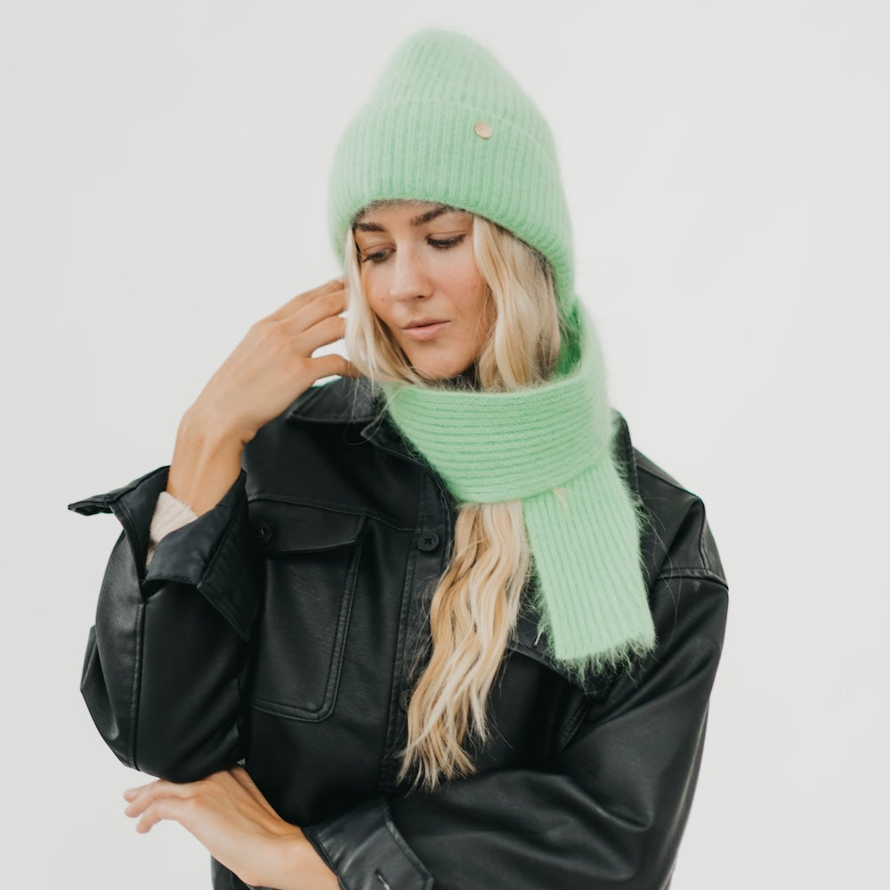 Woman wearing a green knit hat and scarf with a black leather jacket on a light gray background