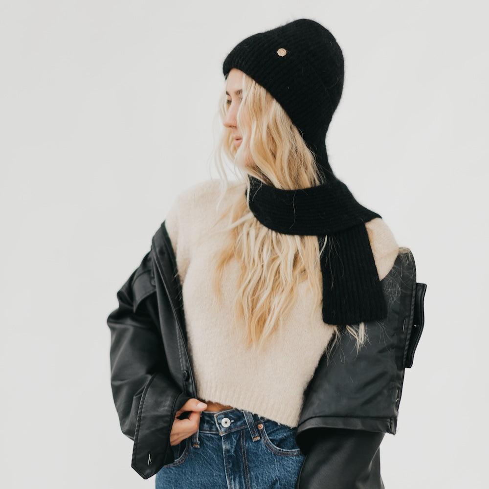 Person wearing a black knit hat and scarf with a leather jacket and jeans on a light gray background