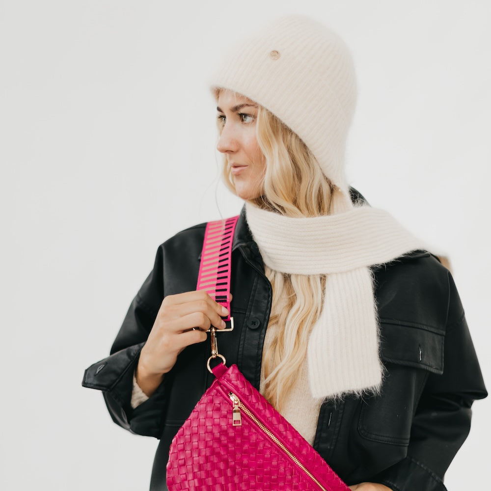 Woman wearing a black coat, cream beanie, and scarf holding a pink bag against a white background