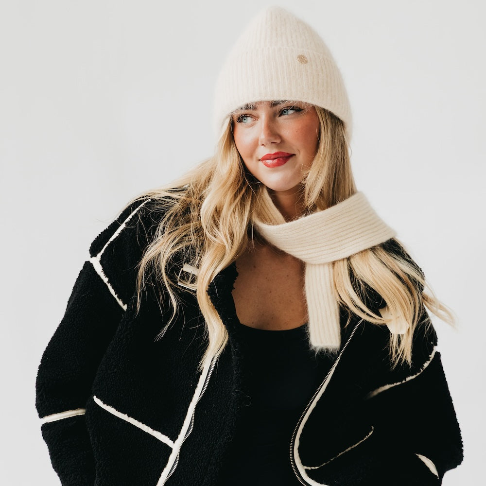 Woman wearing a black coat with white trim, cream beanie, and scarf on a light gray background