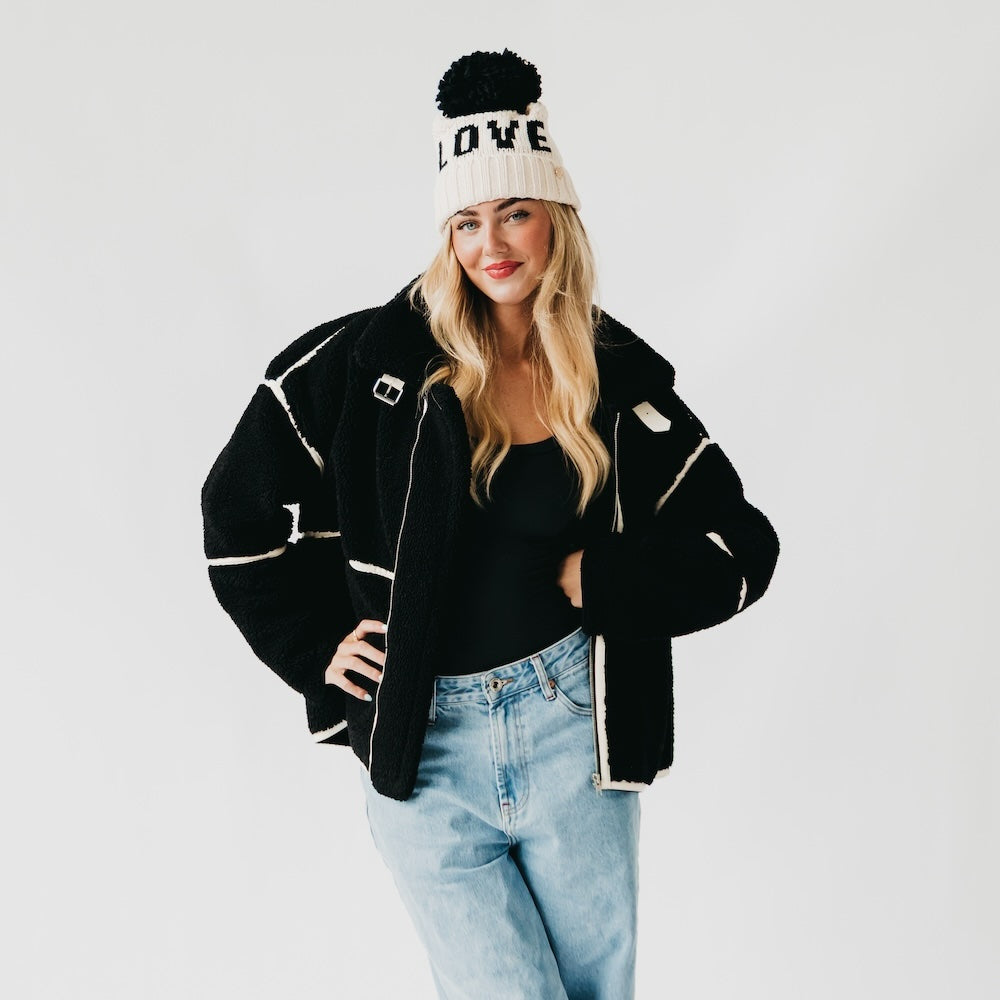 Person wearing a black jacket, black top, and light blue jeans with a white beanie on a plain background