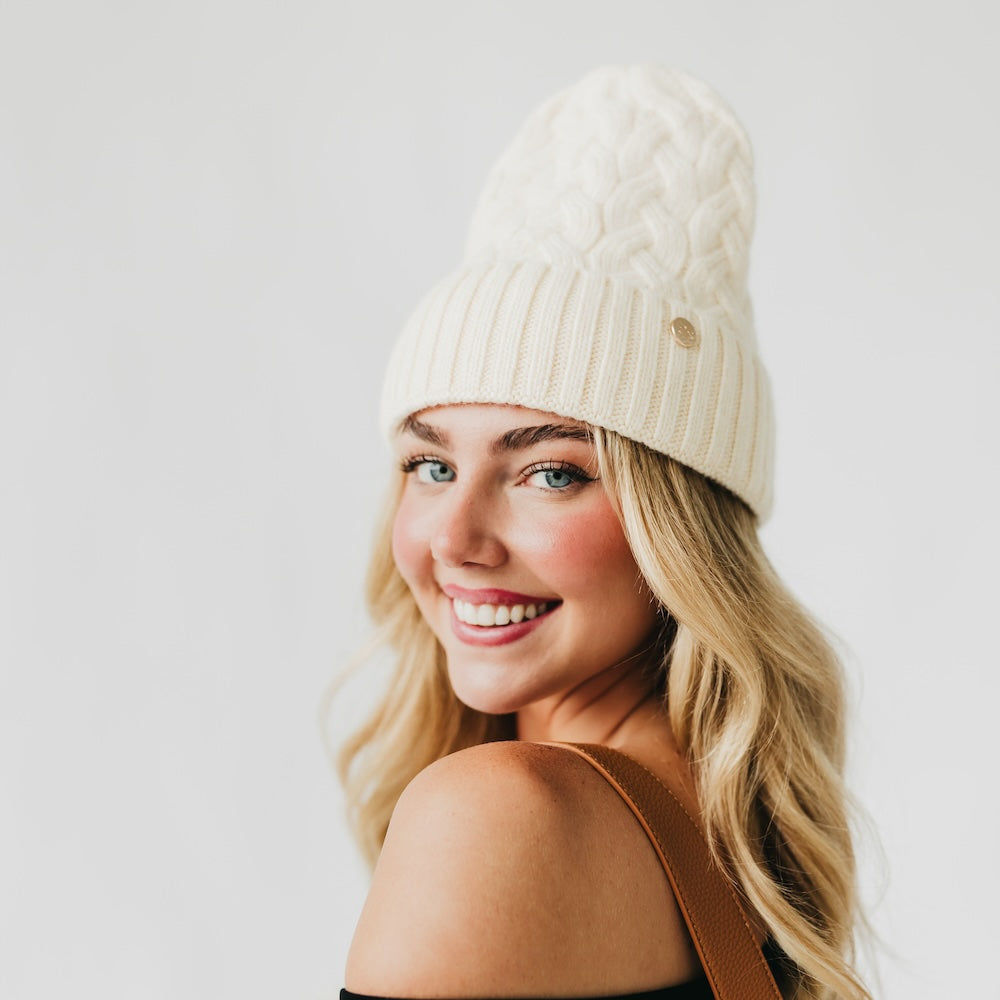 Woman wearing a cream beanie with satin lined interior with a gold logo on a plain background