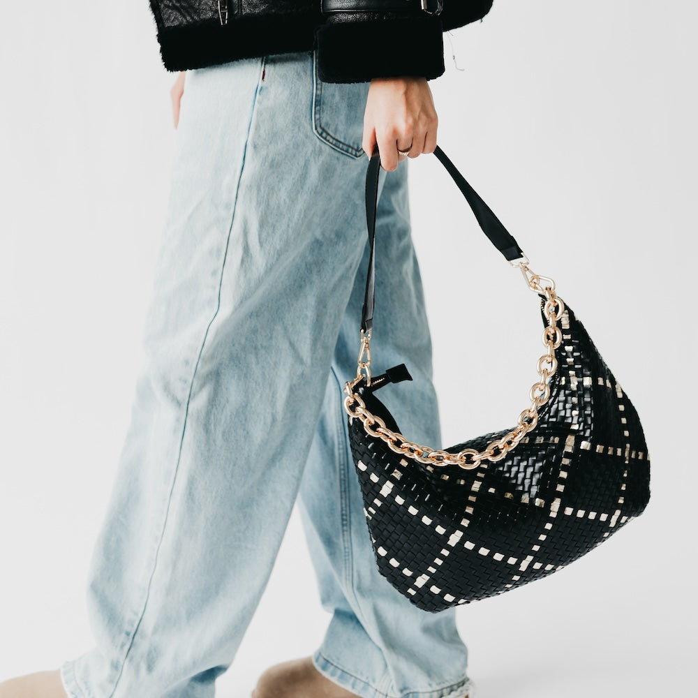 Person holding a black woven handbag with gold chain detail on a plain background