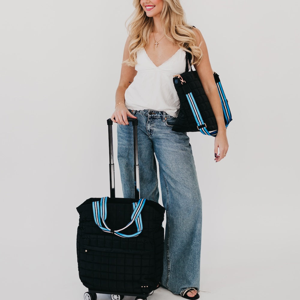 Woman holding a black suitcase and a black tote bag with blue stripes on a plain background