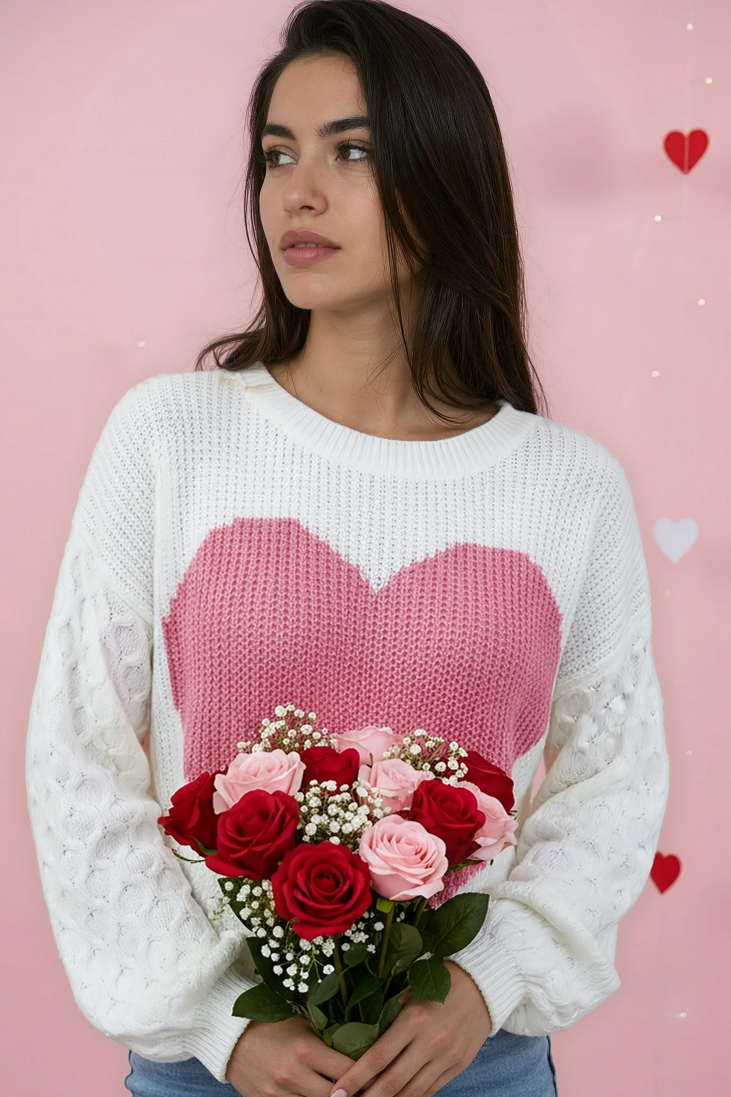 Woman wearing a white sweater with a pink heart design, holding flowers against a pink background.
