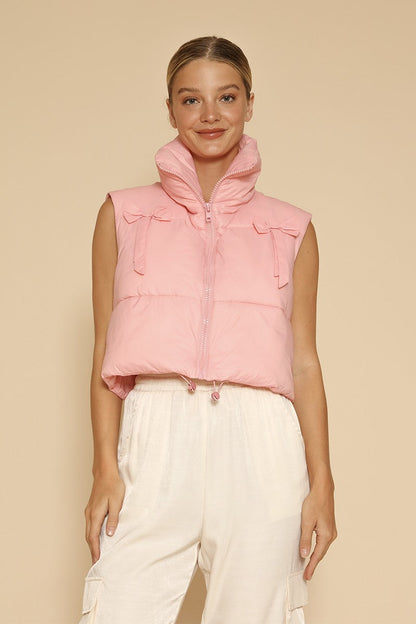 Model wearing a light pink cropped puffer vest featuring front bow detailing near the shoulders, high collar, and zipper closure designed for a feminine yet modern winter look. Girfts for her
