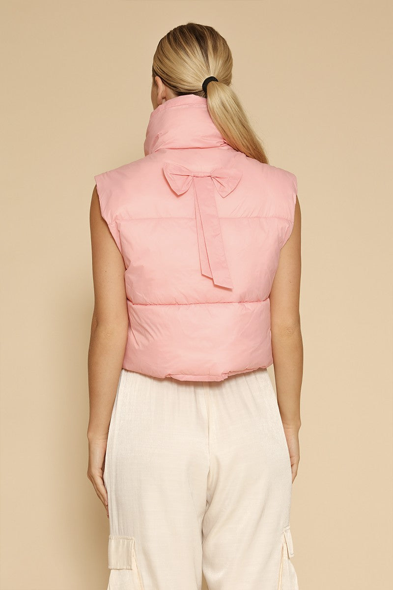 Person wearing a pink puffer vest with a bow on a beige background Back view of a pink cropped puffer vest with a statement bow accent centered below the collar, showcasing its playful and elegant design on a neutral background.