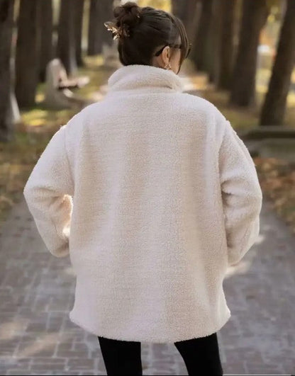 Person wearing a white fluffy coat walking in a park with trees and a path.