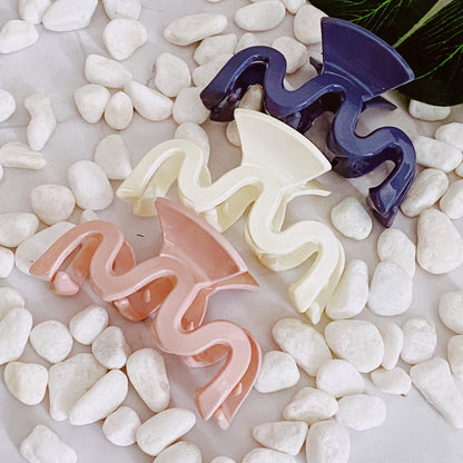 Three wavy ceramic hair clips in purple, white, and pink on pebbles.