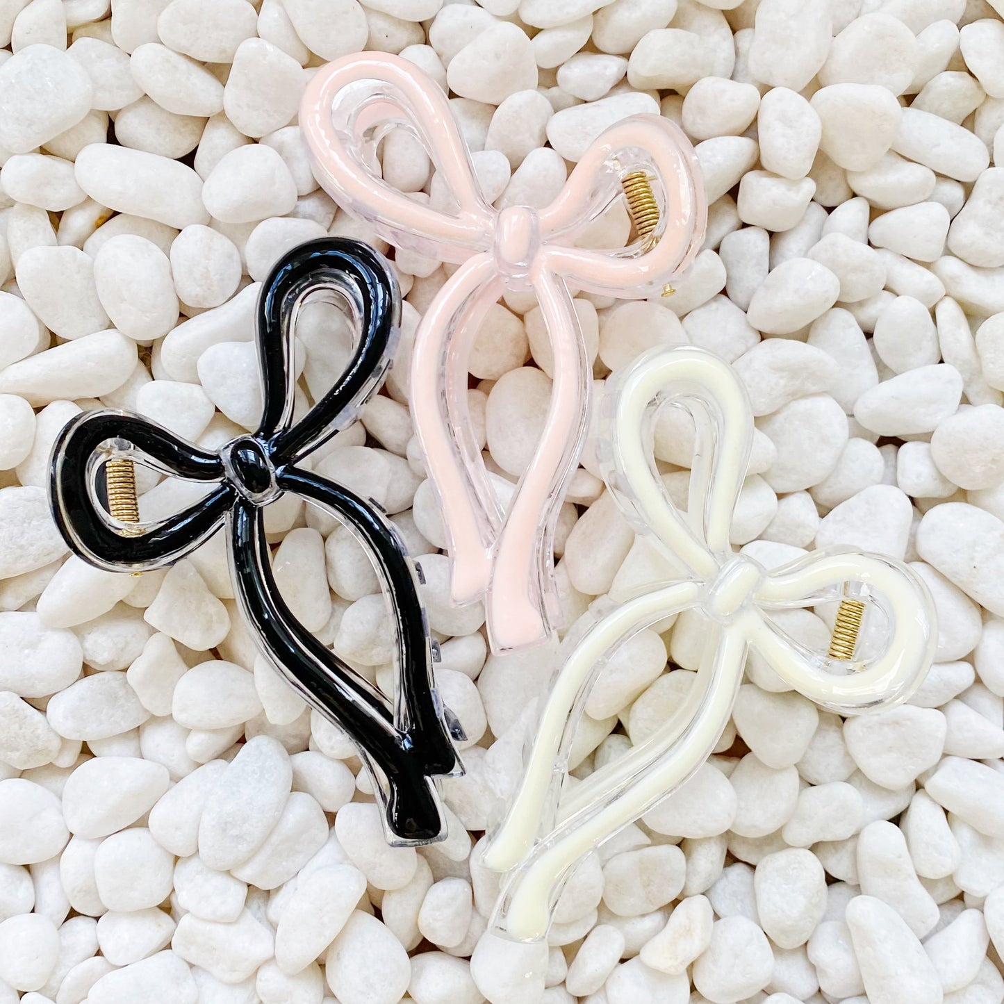 Three bow-shaped hair accessories in black, pink, and white on a pebble background