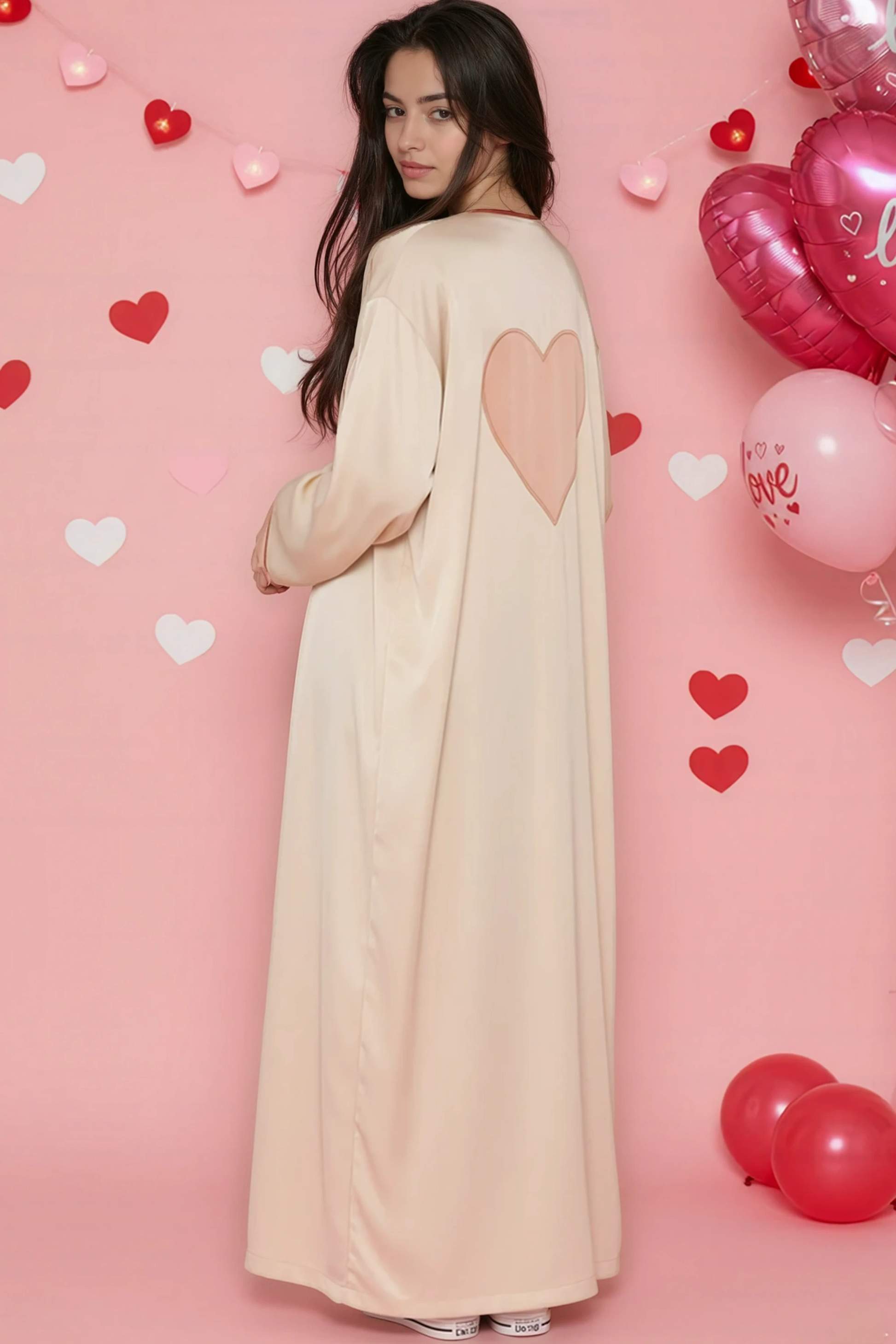 Woman in a long beige dress with heart designs standing against a pink background with heart balloons.