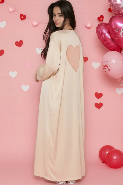 Woman in a long beige dress with heart designs standing against a pink background with heart balloons.
