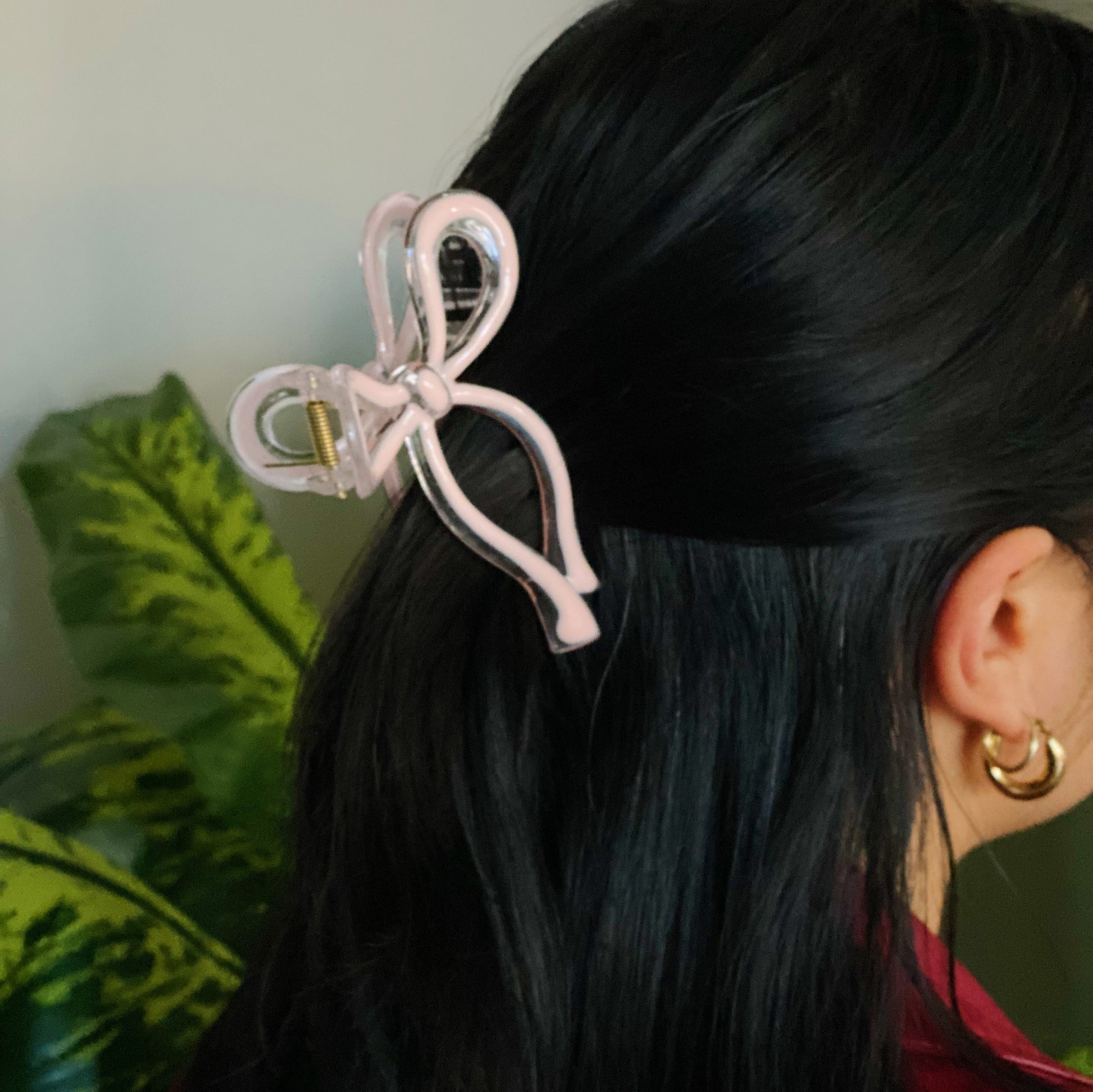 Person wearing a pink hair clip with a bow design in their dark hair, with a plant in the background.