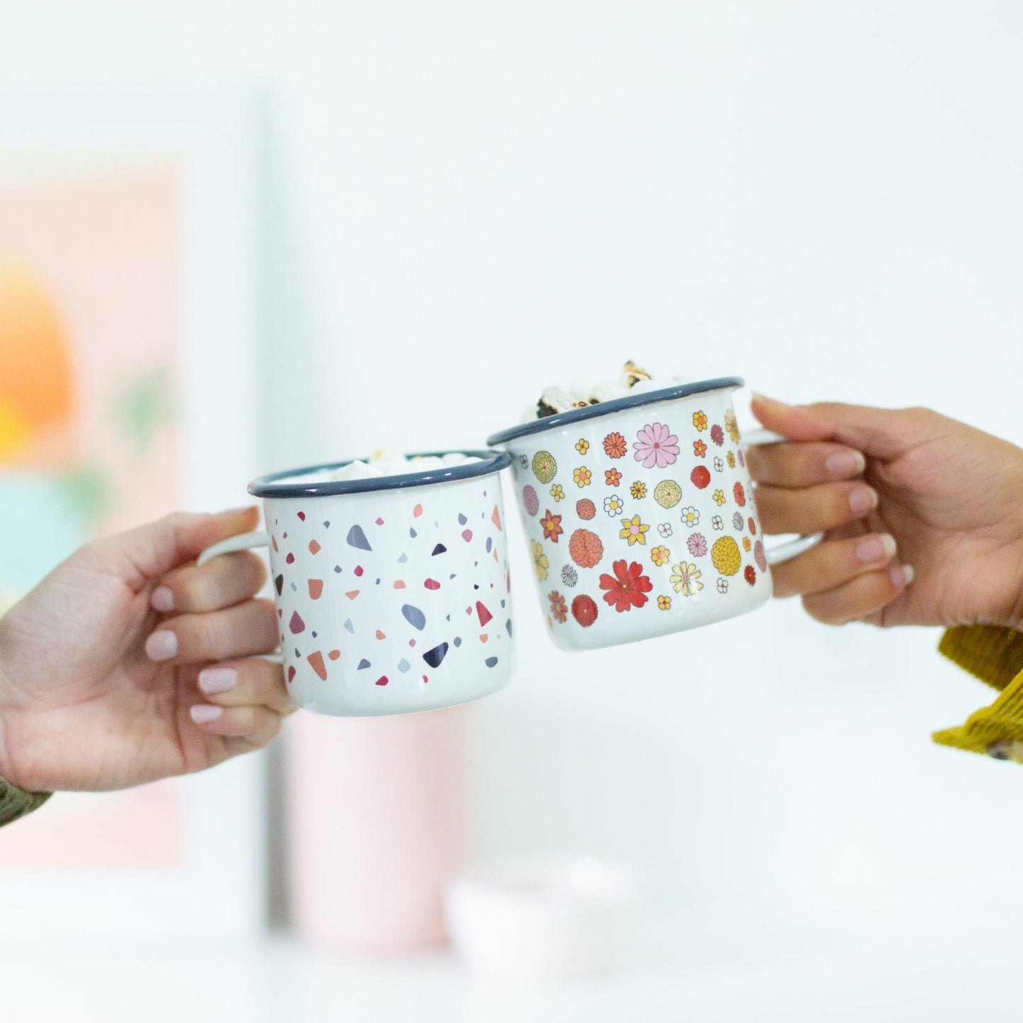 Two hands holding campfire mugs with floral patterns against a blurred background