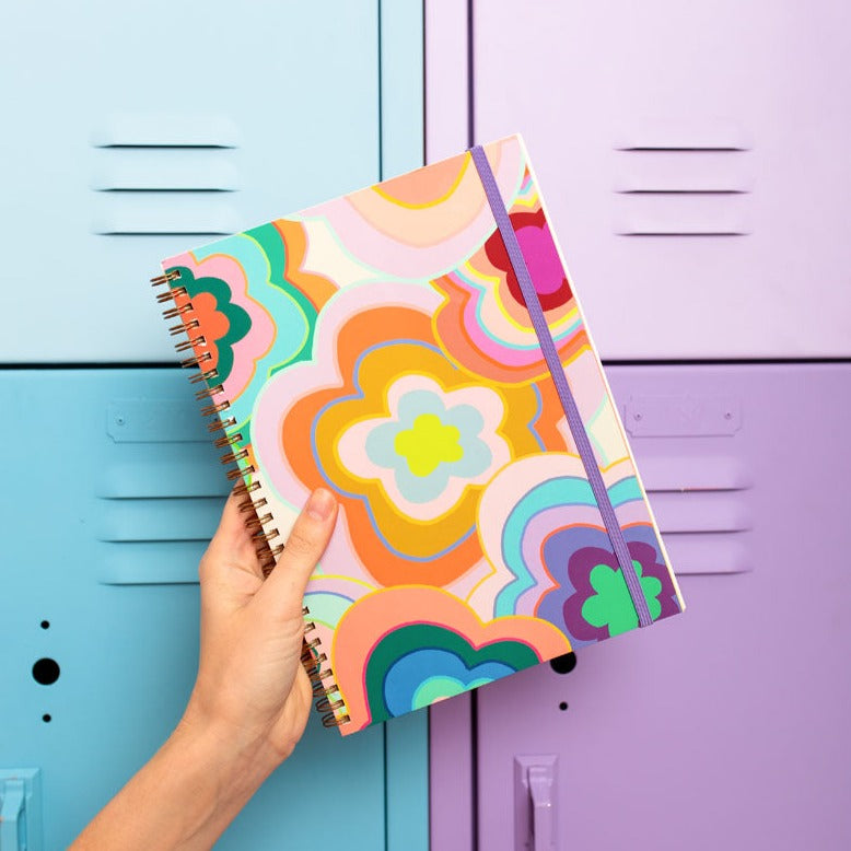 Colorful spiral-bound notebook with a floral design held in front of a blue and purple locker.
