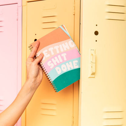 Hand holding a colorful notebook with 'GETTING SHIT DONE' text in front of a yellow locker.