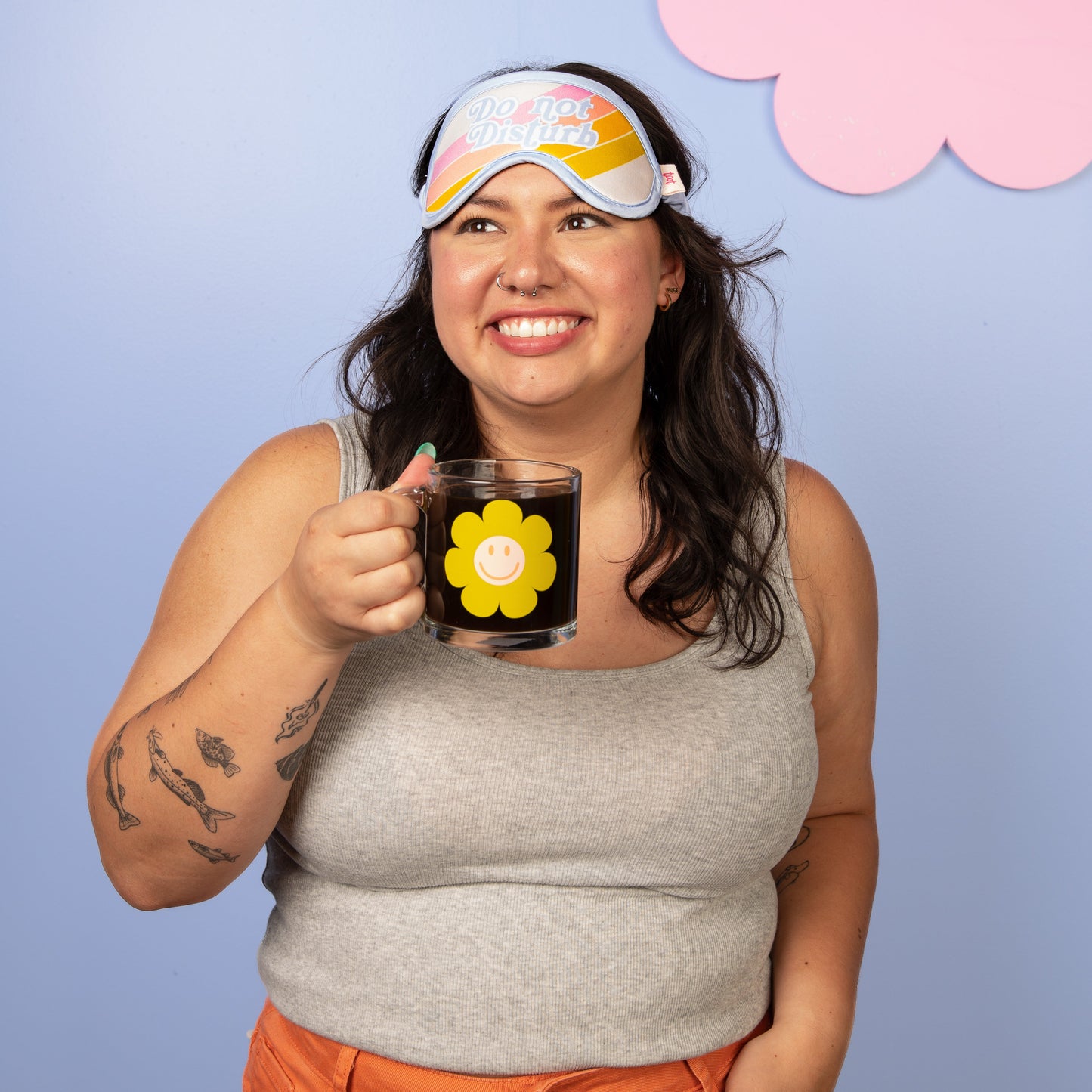 Woman wearing a colorful sleep mask and holding a mug with a flower design against a light blue background.
