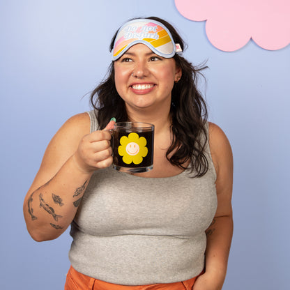 Woman wearing a colorful sleep mask and holding a mug with a flower design against a light blue background.