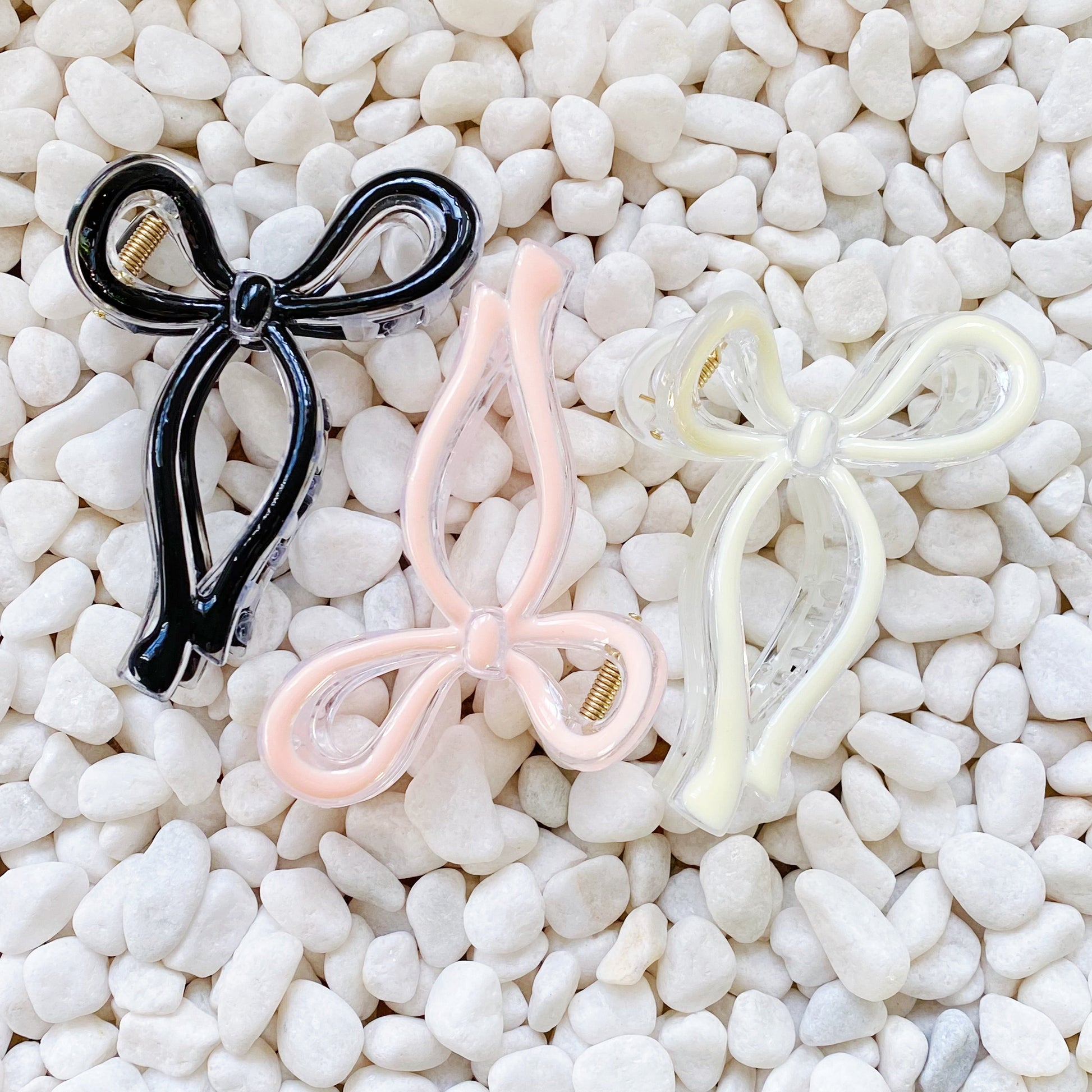 Three bow-shaped hair accessories in black, pink, and white for everyday styling on a pebble background