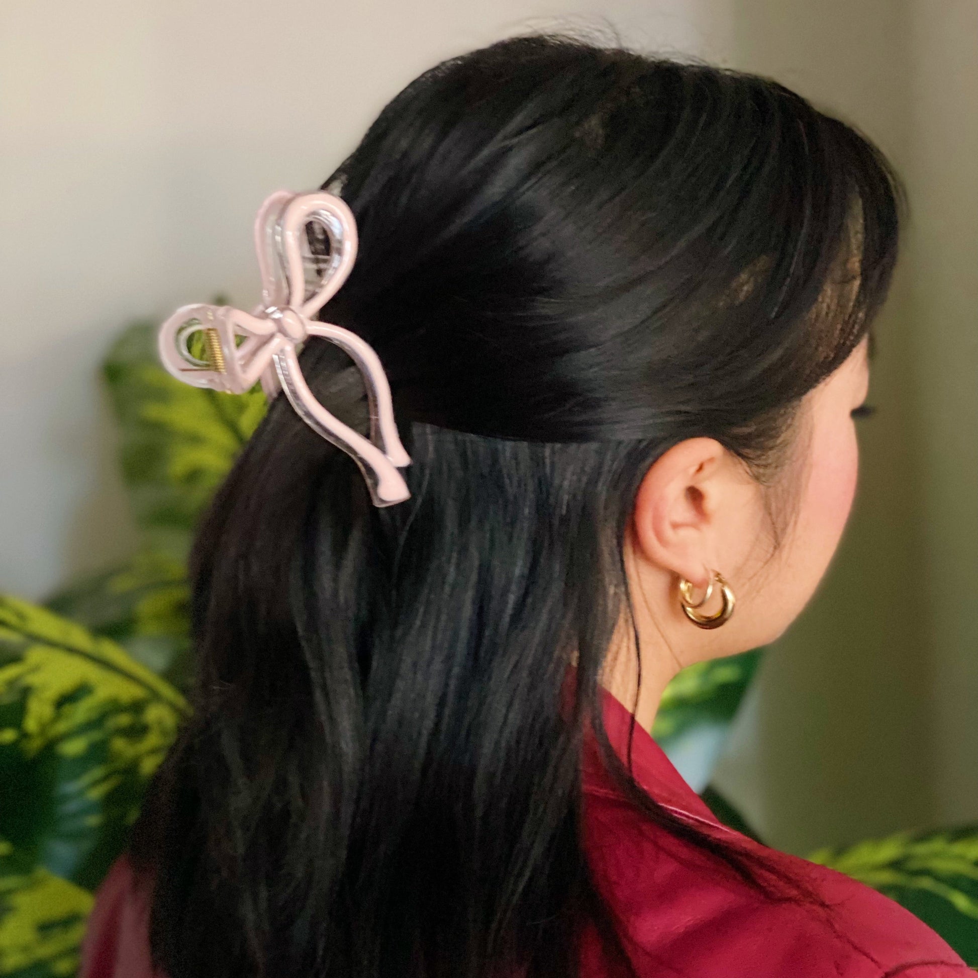 Person with black hair wearing a pink hair bow and gold earring, with a blurred green plant background