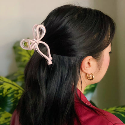 Person with black hair wearing a pink hair bow and gold earring, with a blurred green plant background