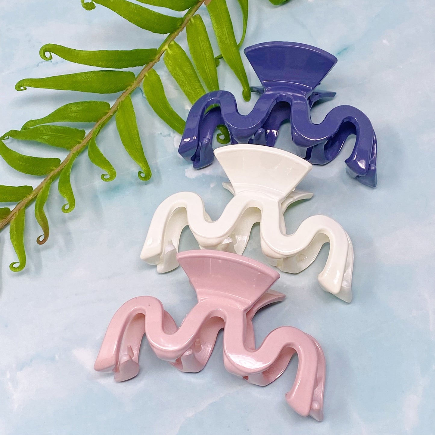 Three colorful wave hair clips (purple, white, pink) on a light blue background with green leaves.