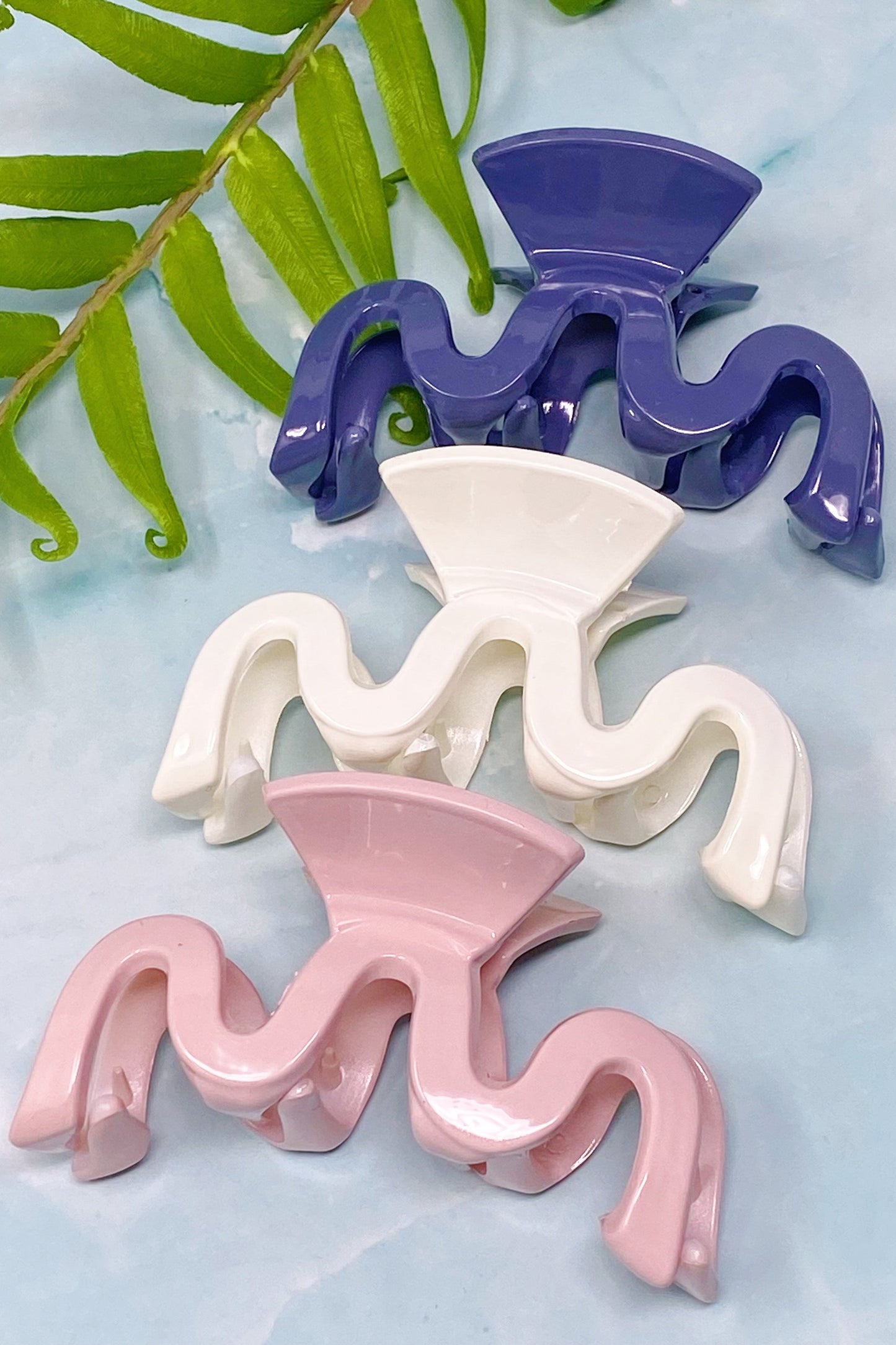 Three wavy wave hair clips in purple, white, and pink on a light blue background with green leaves.