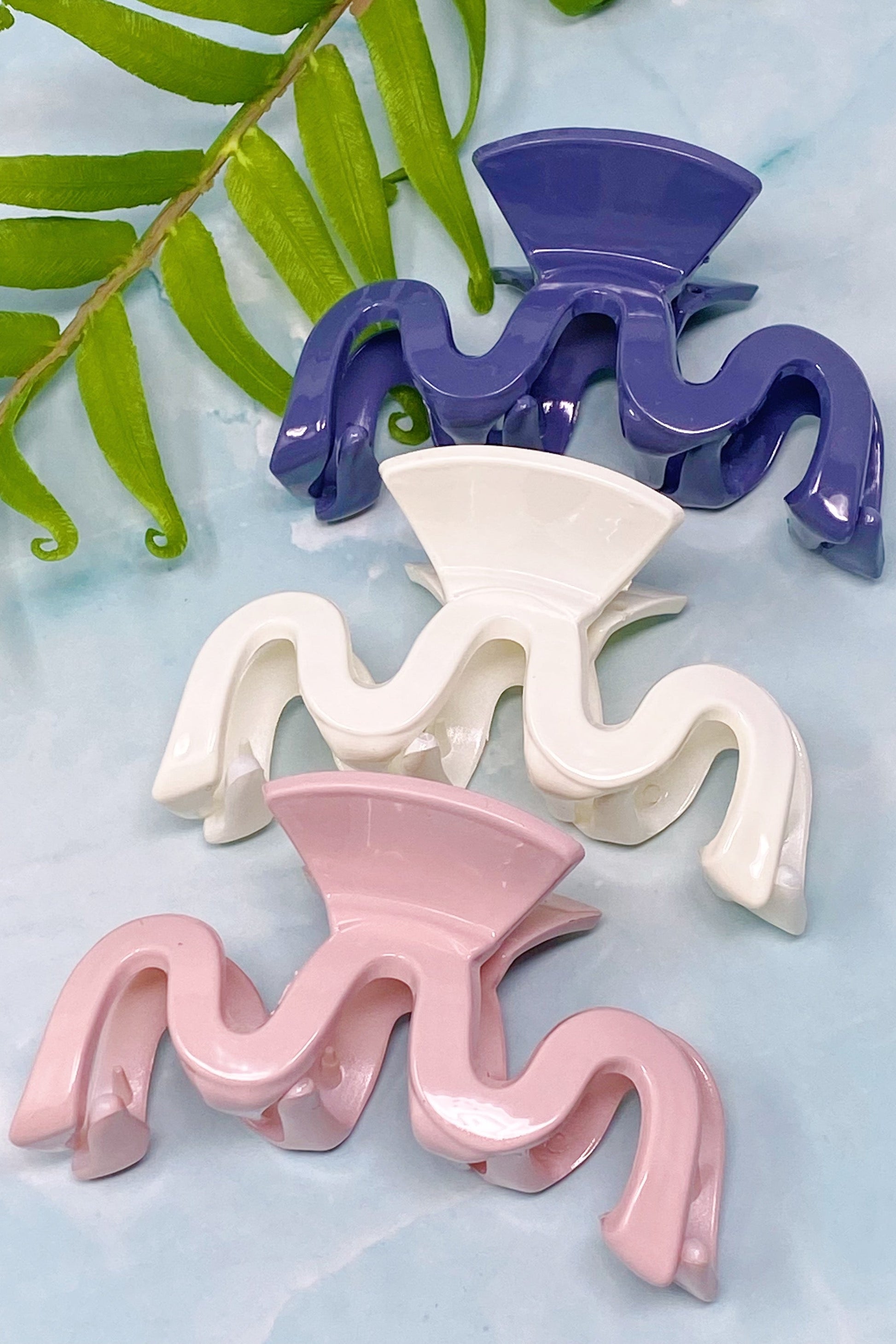 Three wavy wave hair clips in purple, white, and pink on a light blue background with green leaves.