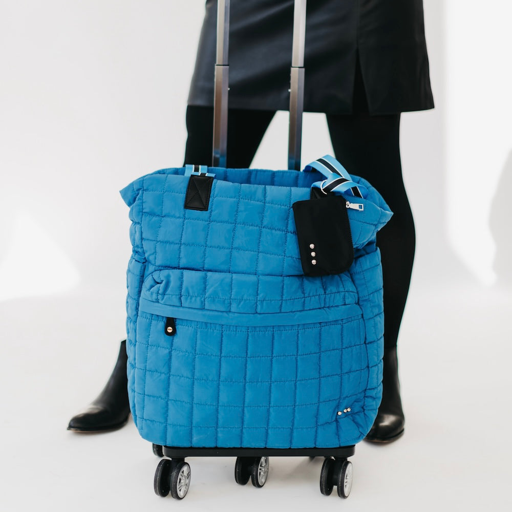 Blue quilted rolling bag with a person wearing black pants and shoes in the background