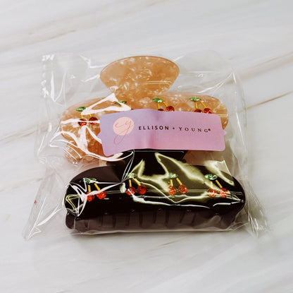 Two hairclips with cherry design in clear packaging with a visible brand label on a light surface.