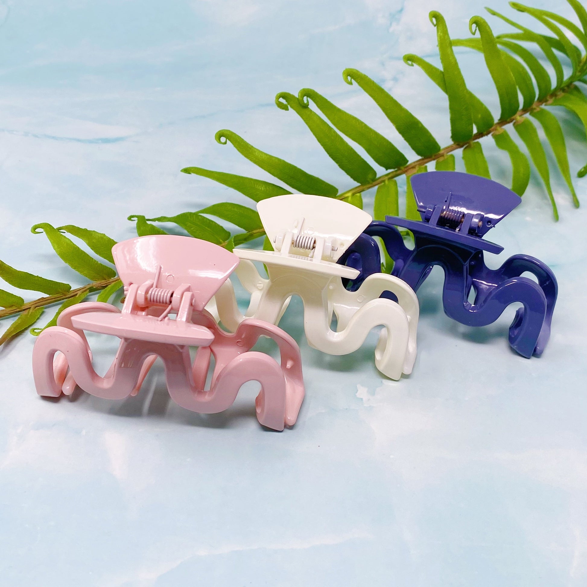 Three wave hair clips in pink, white, and blue on a light blue background with green leaves.