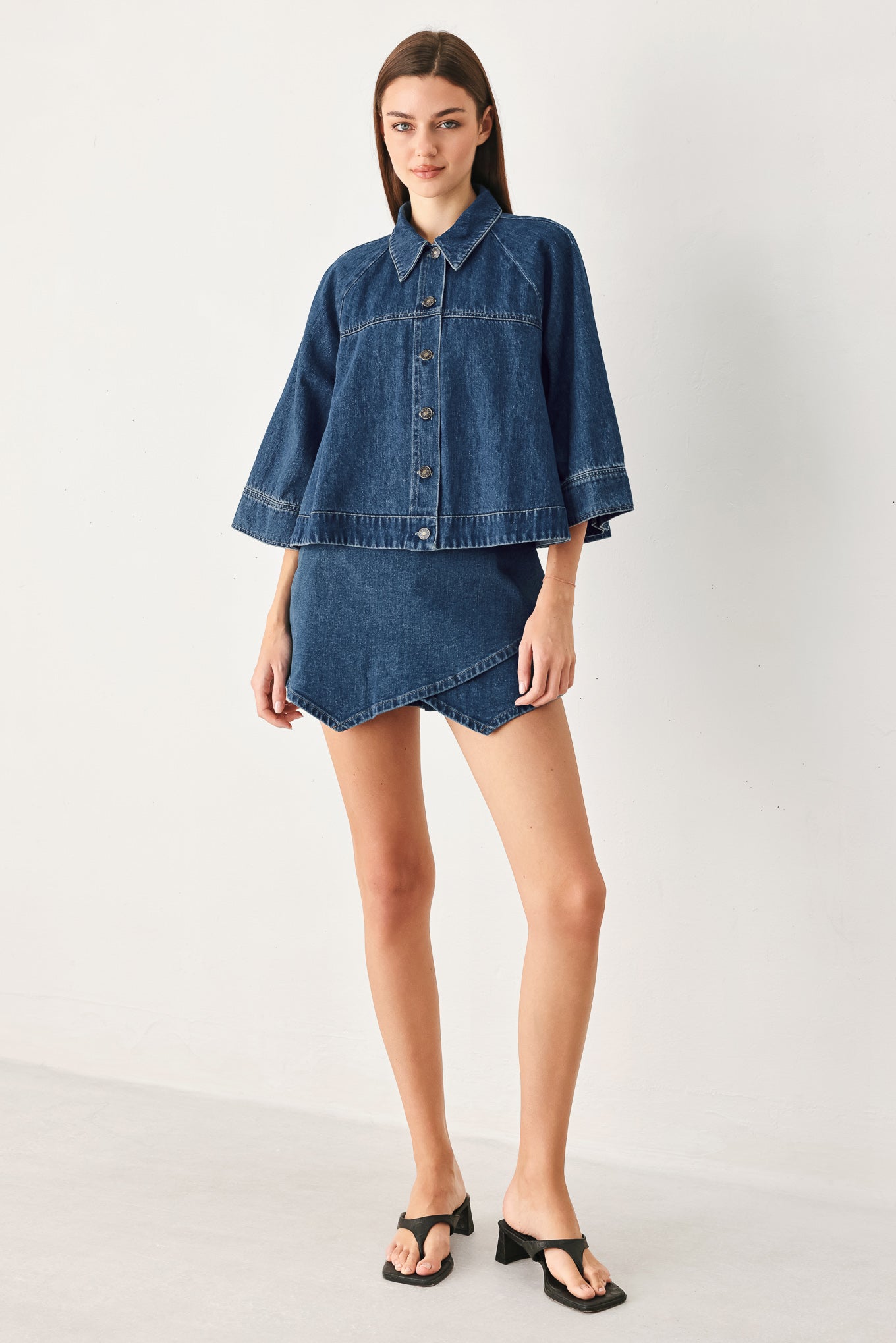 A model wearing a blue denim short-sleeve button-up shirt and matching denim mini skirt, paired with black thong sandals, standing against a plain light gray studio background.