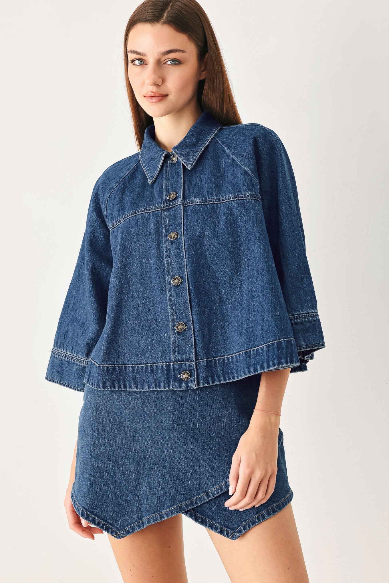 A model wearing a blue denim jacket with a matching denim mini skirt, photographed against a plain light gray studio background.