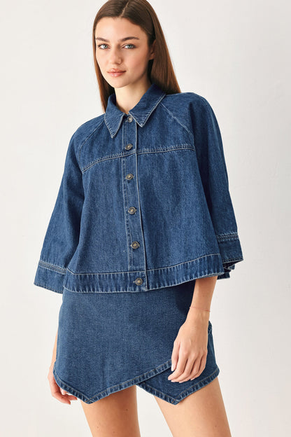 A model wearing a blue denim jacket with a matching denim mini skirt, photographed against a plain light gray studio background.