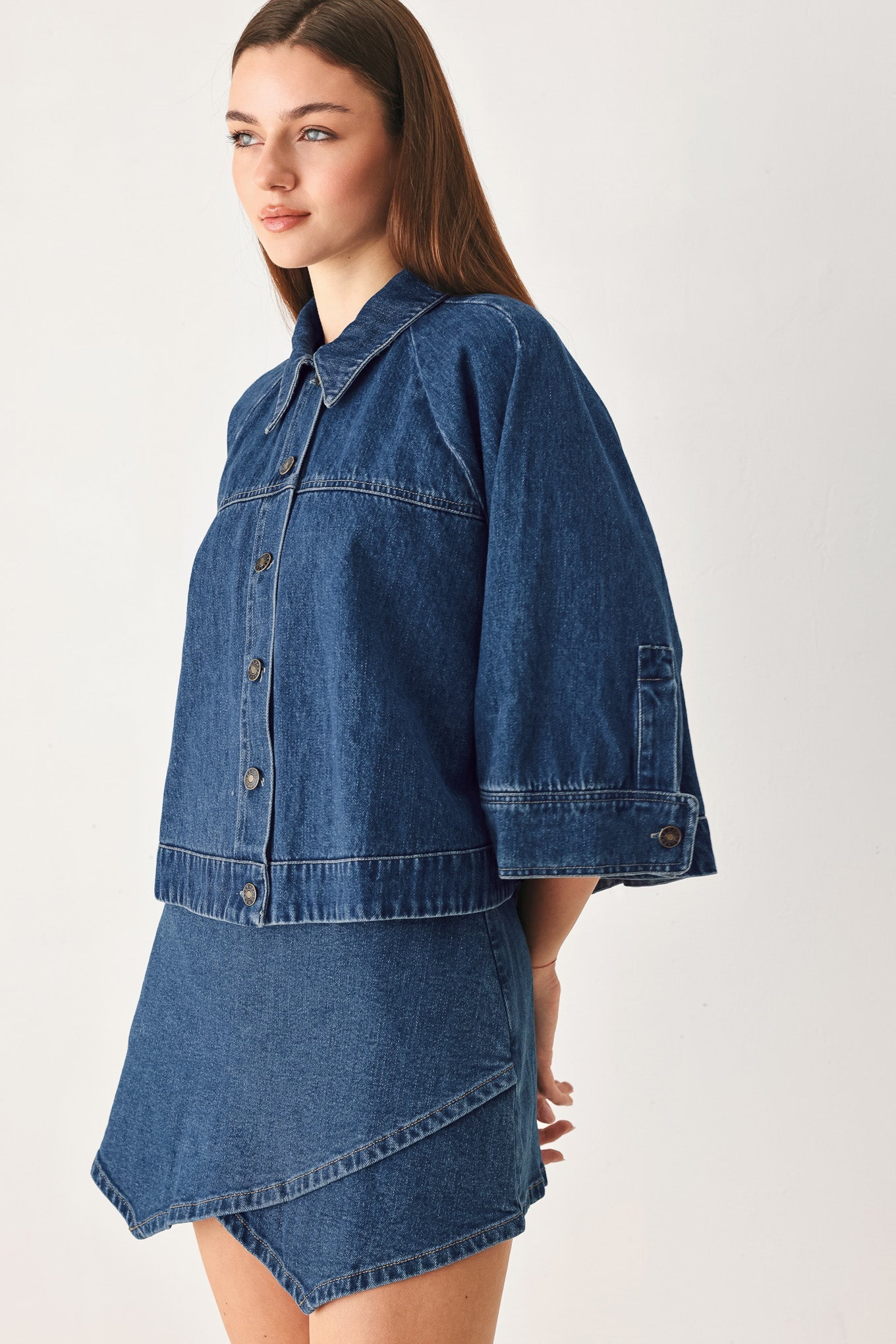 A woman wearing a blue denim shirt-jacket and matching denim skirt, photographed against a plain light gray studio background.