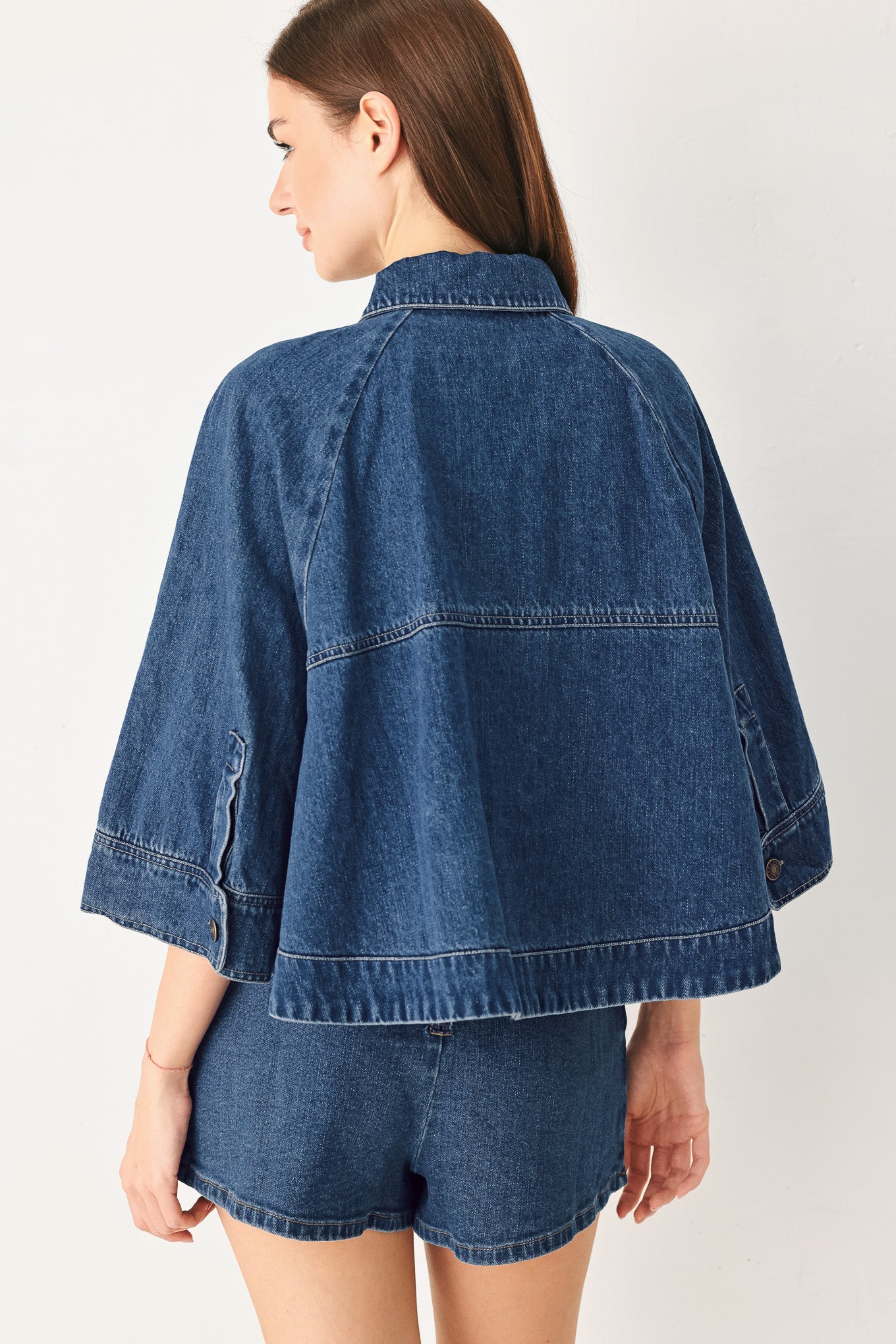 A woman shown from the back wearing a cropped blue denim jacket with wide three-quarter sleeves and matching denim shorts, against a plain light background.