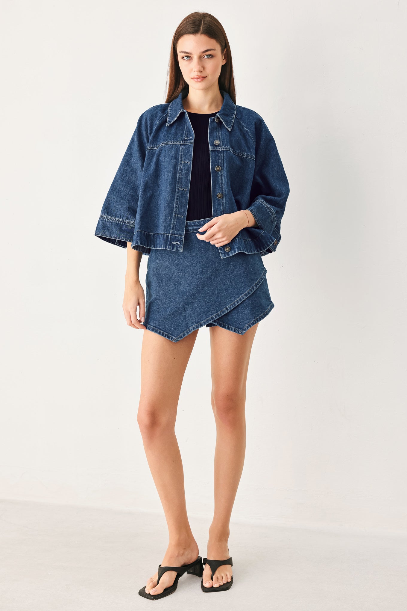A model stands against a plain light gray studio background wearing a blue denim cropped jacket with wide sleeves, a matching blue denim mini skirt, a black top, and black thong sandals.