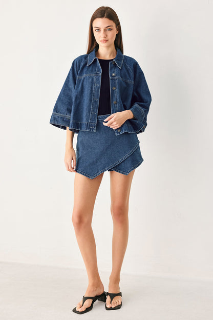 A model stands against a plain light gray studio background wearing a blue denim cropped jacket with wide sleeves, a matching blue denim mini skirt, a black top, and black thong sandals.